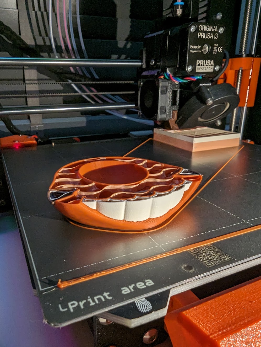 Picture shows an undergoing multi-material print. You can see the layers with different colors, and a tower in the back, where the extruder is wiping old filament in order to change colors